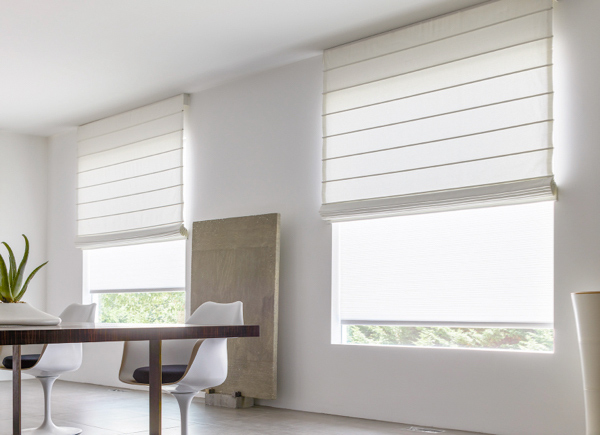 Two large windows featuring Cascade Roman Shades in a dining room with a large wooden plank art and table with white chairs