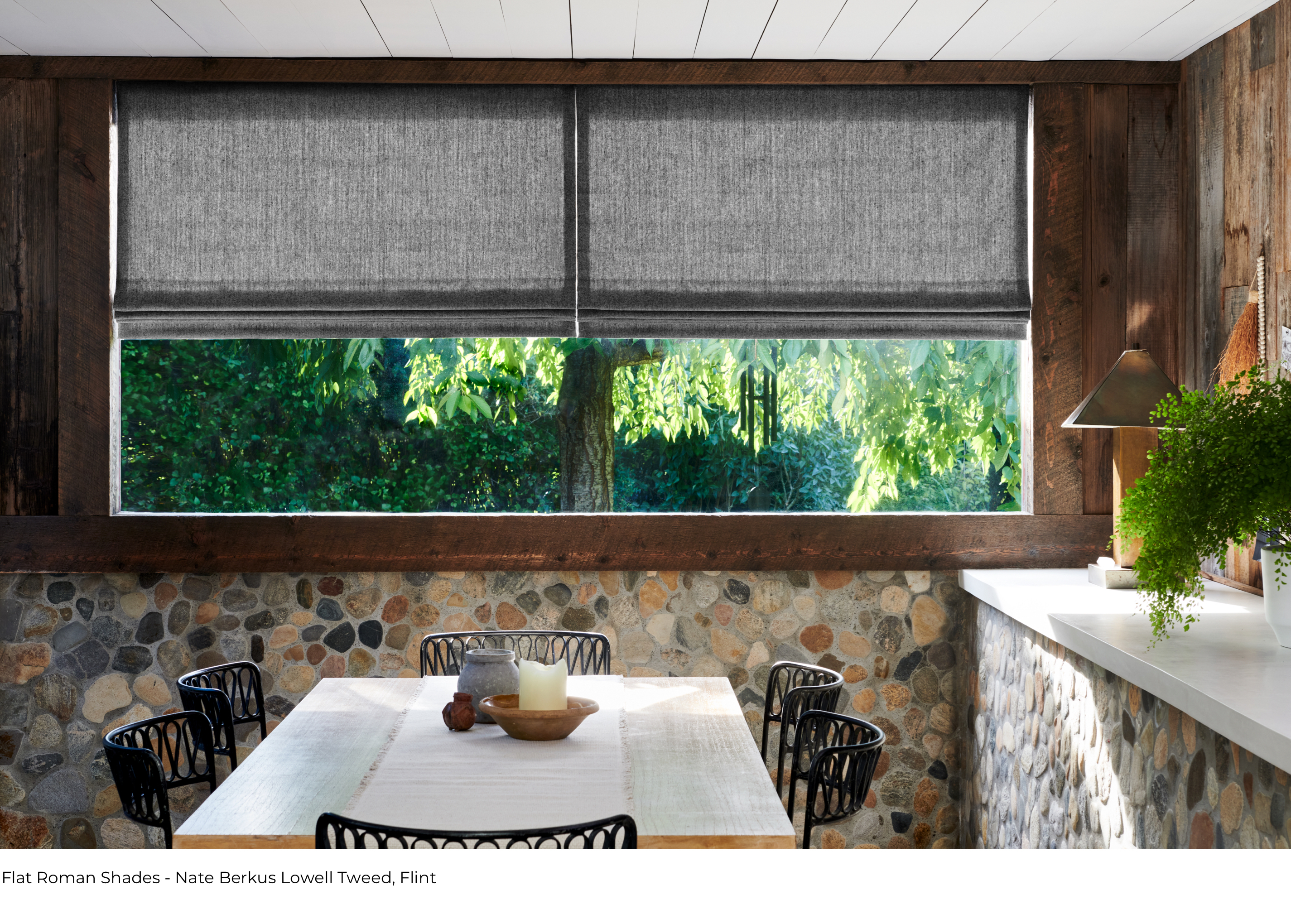 A large window featuring Flat Roman Shades in Lowell Tweed Flint in a dining room with a half rock wall and a wood table