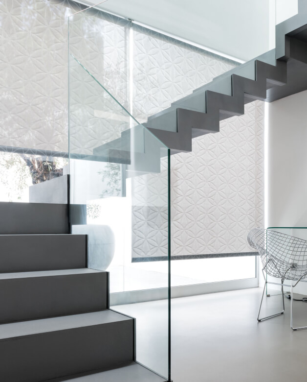 A bright, modern stairwell with a glass railing and patterned motorized Solar Shades in a neutral colorway.