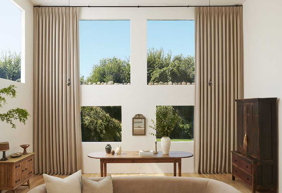 Pinch Pleat Drapery frames a convivial living room scene with plenty of greenery outside and a couch in the foreground, offering curtain inspiration for tall ceilings and windows