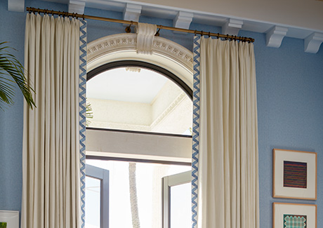 A blue room with a tall ceiling features curtains on arched windows made of Tailored Pleat Drapery in Luxe Linen, Oyster