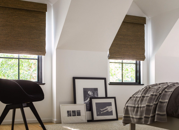Two windows featuring woven wood shades in a bedroom with dark colored bedding and framed art placed against the wall