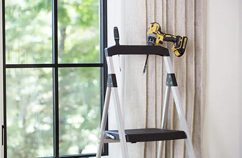 A step ladder and drill in front of a window show some of the tools needed to learn how to install roman blinds