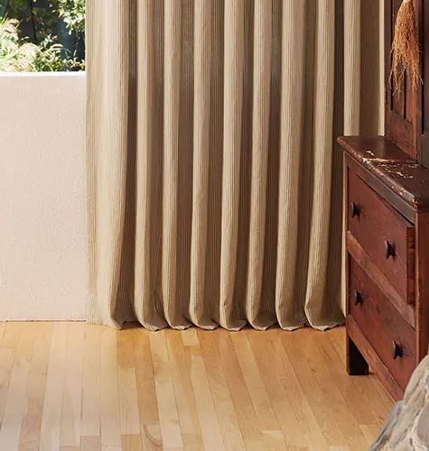 Pinch Pleat Drapery in Surrey Stripe Morel and Natural beside a wood chest, showing floor length curtains touching the floor with a tailored break length design.